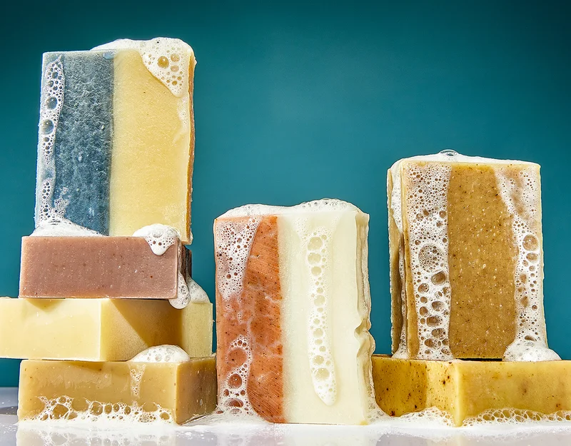 Six bars of sudsy soap are piled onto each other. They come in different colors (earth tones). All against a dark teal blue background.
