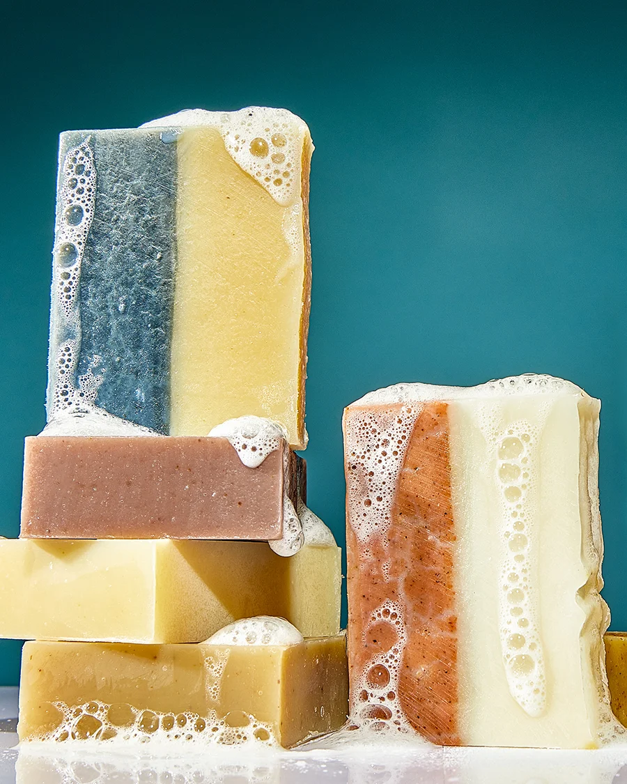 Six bars of sudsy soap are piled onto each other. They come in different colors (earth tones). All against a dark teal blue background.