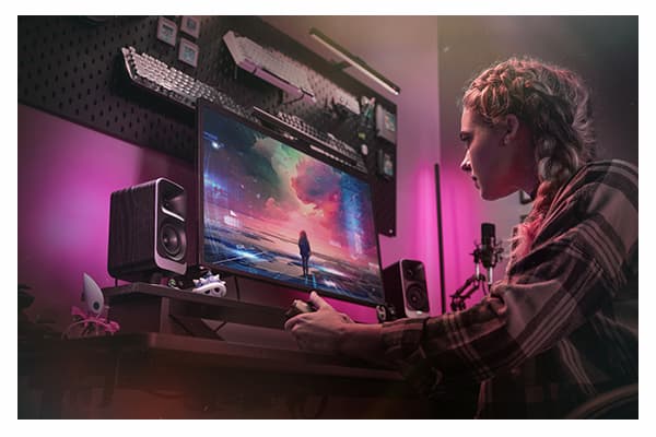 ProMedia Lumina Computer Speakers with LED Lighting. Young woman playing a game on her computer with ProMedia Lumina Speakers on either side. Speakers project pink LED light on the wall behind them.