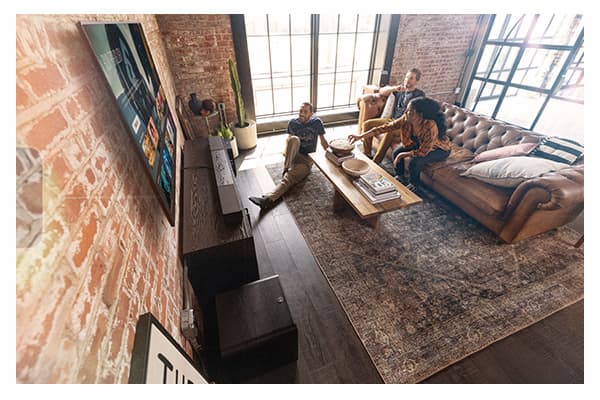 Flexus CORE 200 3.1.2-Channel Dolby Atmos Sound Bar. Two men and a woman with popcorn, watching a flat-screen tv, hanging on a brick wall, in a rustic modern living room.  A black sound bar sits on a black cabinet just below the television.