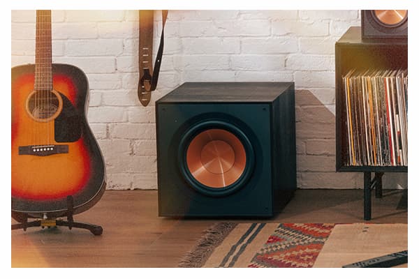 R-121SW Subwoofer. Subwoofer is sitting on a wooden floor, in front of a white brick wall. While also surrounded by a guitar, a cabinet full of vinyl records, and an ornate knit rug.