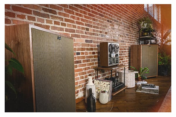 Cornwall IV Floorstanding Speaker. Pair of walnut finished floor standing speakers seated against the brick wall of a living room.  A wooden stand with a tape-deck, records, vases and a basket is situated between the speakers. Further along the wall, an behind the speaker are shelving units with house plants