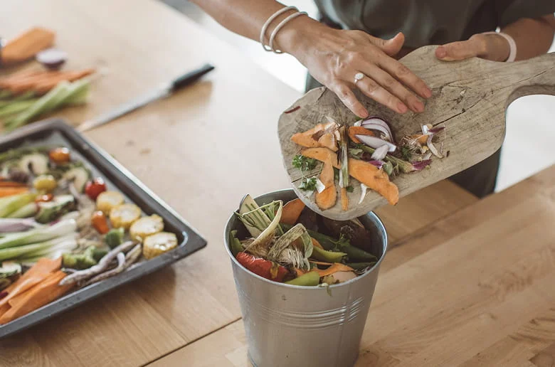 Someone composts vegetable scraps from a cutting board into a bin, with a tray of colorful chopped veggies nearby, highlighting sustainable kitchen habits.