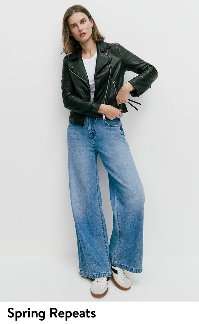 A woman wearing a black moto jacket with a white T-shirt, wide-leg jeans and sneakers.