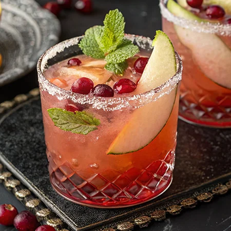 A cocktail in a textured glass features red liquid with ice, apple slices, cranberries, mint leaves, and a sugar rim.