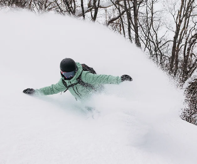 Snowboarder wearing a yellow jacket and blue snowboard pants rides through deep powder, spraying snow, with a white snow-covered mountain slope in the background.