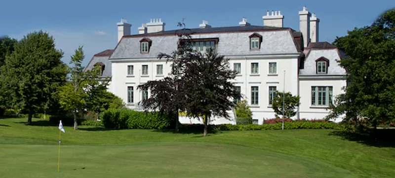 Grand white estate facing a golf green, with manicured lawn, trees, and clubhouse-style architecture.