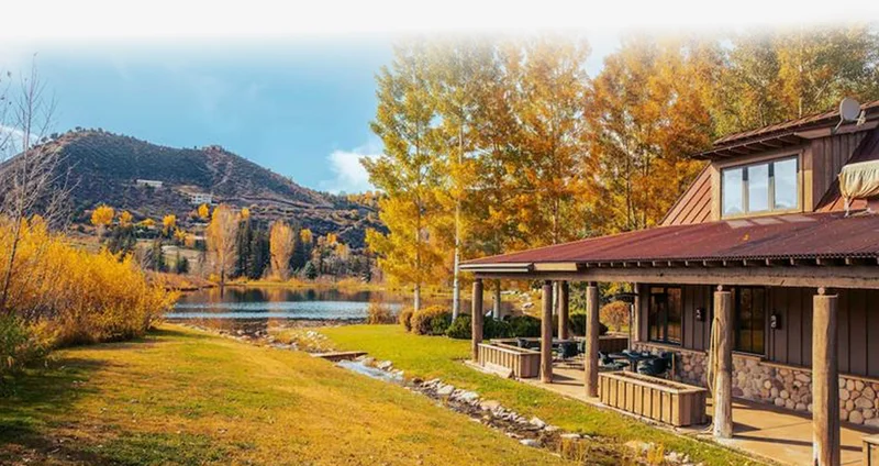 Lakeside cabin with covered porch and outdoor seating, framed by autumn trees and water views.