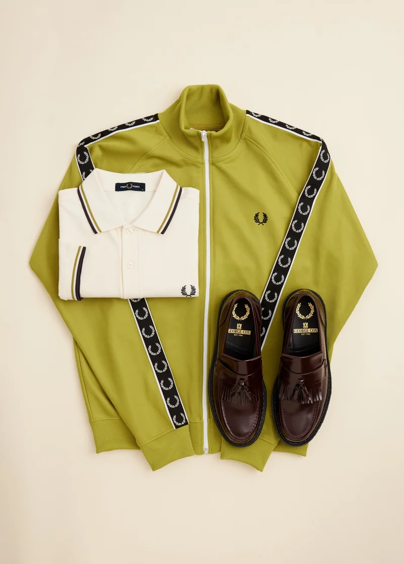The Gift List: For Him. Image shows a flat lay image of a lime greenish yellow track jacket, a folded twin-tipped ecru Fred Perry shirt and a pair of George Cox loafers.