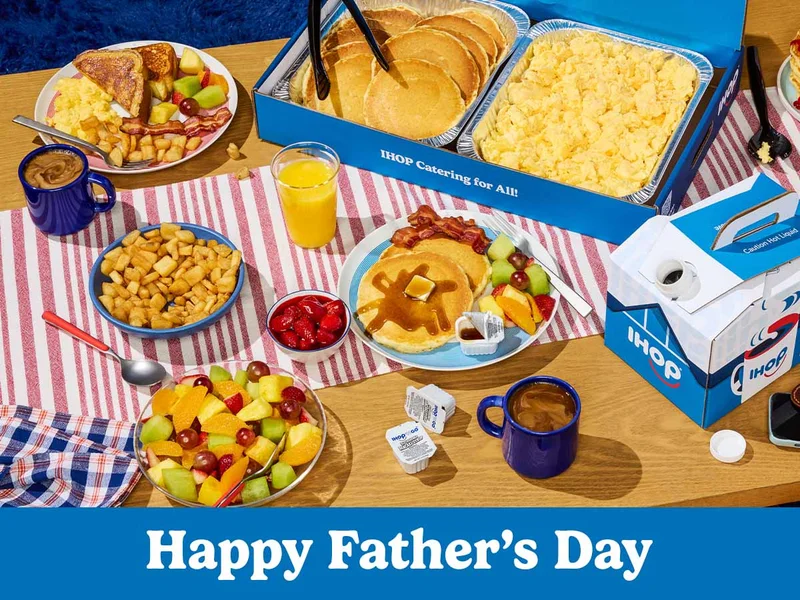 An arrray of IHOP catering on a wood table with a red and white striped runner. Food includes tins of pancakes and scrambled eggs, a box of coffee, and a bowl of fresh fruit