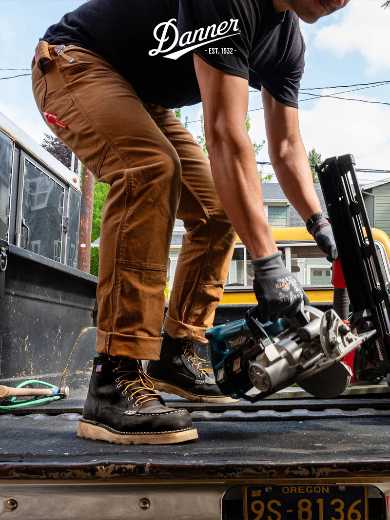 A person in brown pants and a black t-shirt is stepping onto the tailgate of a pickup truck, wearing black Danner Bull Run Moc Toe boots. They are grabbing a circular saw near other tools and building materials.