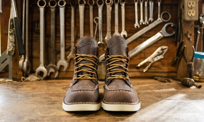 A pair of brown Danner Bull Run Moc Toe boots facing forward on a wooden workbench, with an array of tools like wrenches and pliers hanging on a wooden wall in the background.