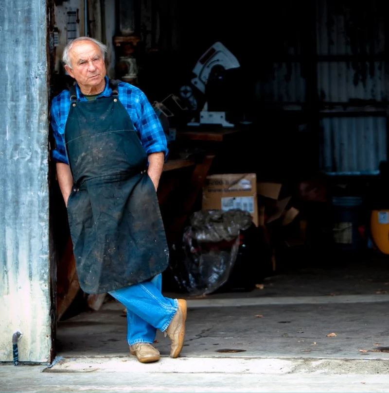 A person in a dirty apron stands in the doorway of a blacksmith shop.