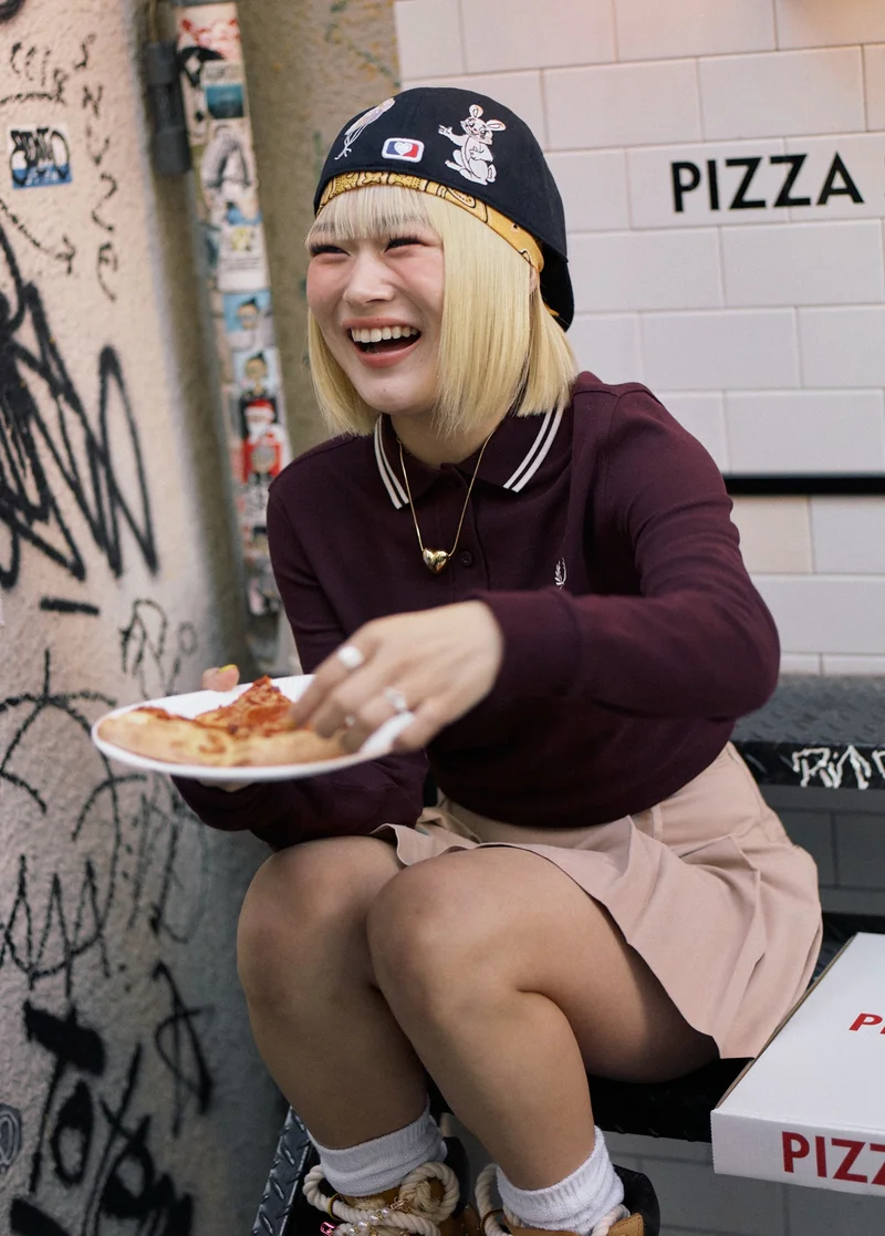 Female Asian model bleached hair in a bob cut with a fringe sits on a step outside a pizzeria and is giggling while picking up a slice of pizza. She wears a twin-tipped Oxbloof Fred Perry Shirt, a peach-coloured miniskirt and an embroidered backwards cap.