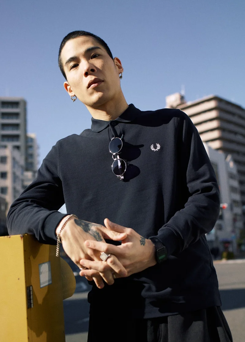 Asian male model with buzzcut leans on a metal locker on a city street with his hands interlocked. He wears a long sleeve black Fred Perry Shirt which is accessorised with hoop earrings, a chain bracelet, a signet ring and a pair of sunglasses hanging from the collar.