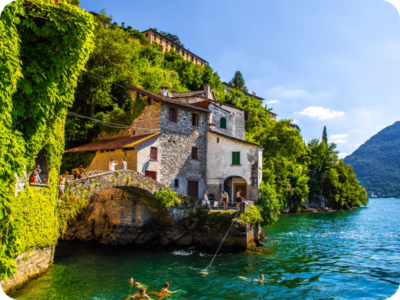 People swim by a stone bridge and house in Lake Como, Italy