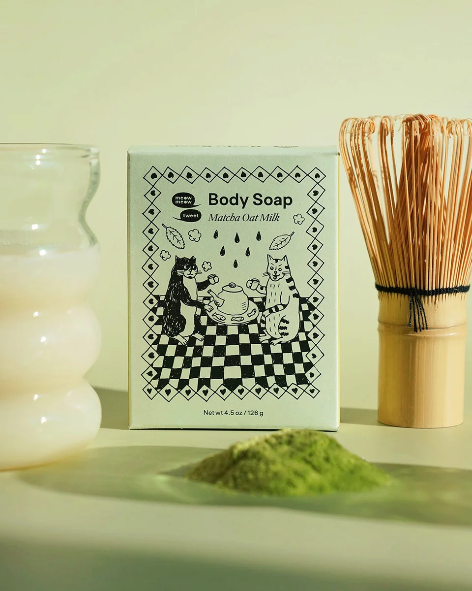 A light green box with a pile of green matcha powder in front of it. A matcha brush and a glass of milk set to each side.