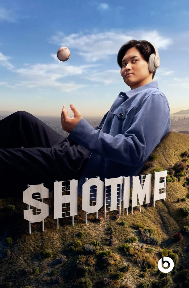 Baseball player Shohei Ohtani, wearing Beats Studio Pro headphones, sits on a hillside with a baseball floating above his hands, in front of large