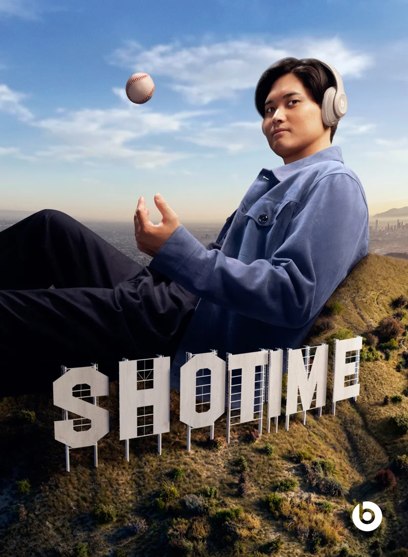 Baseball player Shohei Ohtani, wearing Beats Studio Pro headphones, sits on a hillside with a baseball floating above his hands, in front of large