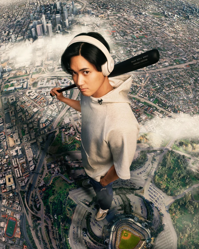 Shohei Ohtani, wearing Beats Studio Pro headphones and a hoodie and holding a baseball bat, stands towering over a miniature cityscape with a serious expression.