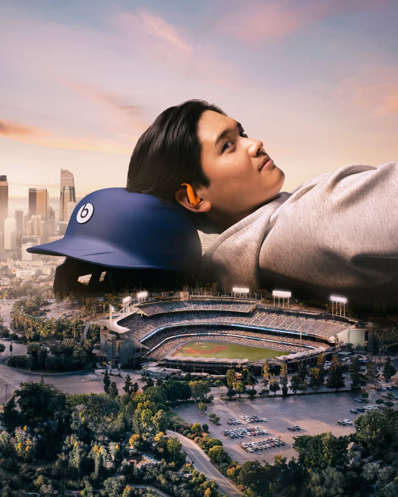 Shohei Ohtani, wearing orange Powerbeats Pro 2 earbuds, reclines with a blue baseball helmet as a pillow, looking at a miniature city and a bustling baseball stadium below him.