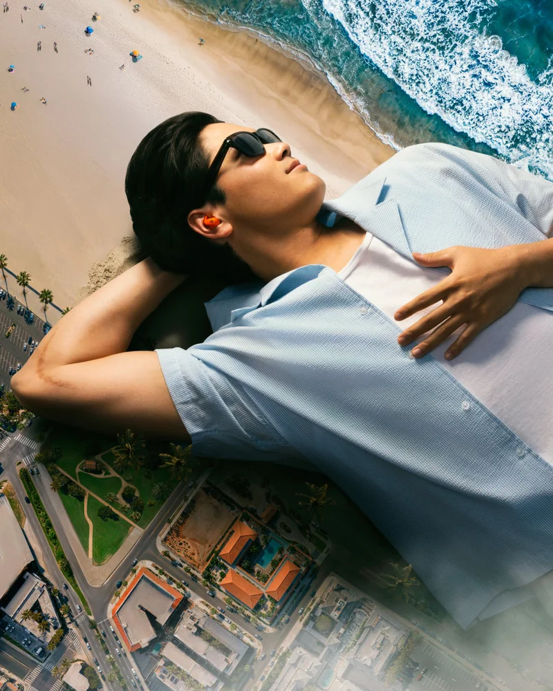 Shohei Ohtani, wearing sunglasses and orange Powerbeats Fit earbuds, lies relaxed on a sandy beach, looking up, with a miniature city and ocean spread out beneath him.