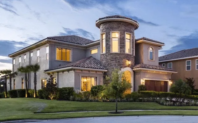 Two-story Mediterranean-style home with stone tower and warm exterior lighting at dusk.