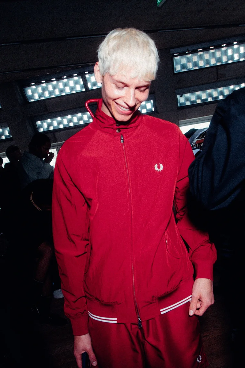 Male white model with short bleached hair wears the red crinkle-textured Harrington Jacket backstage. He is looking down and smiling as the camera flashes.