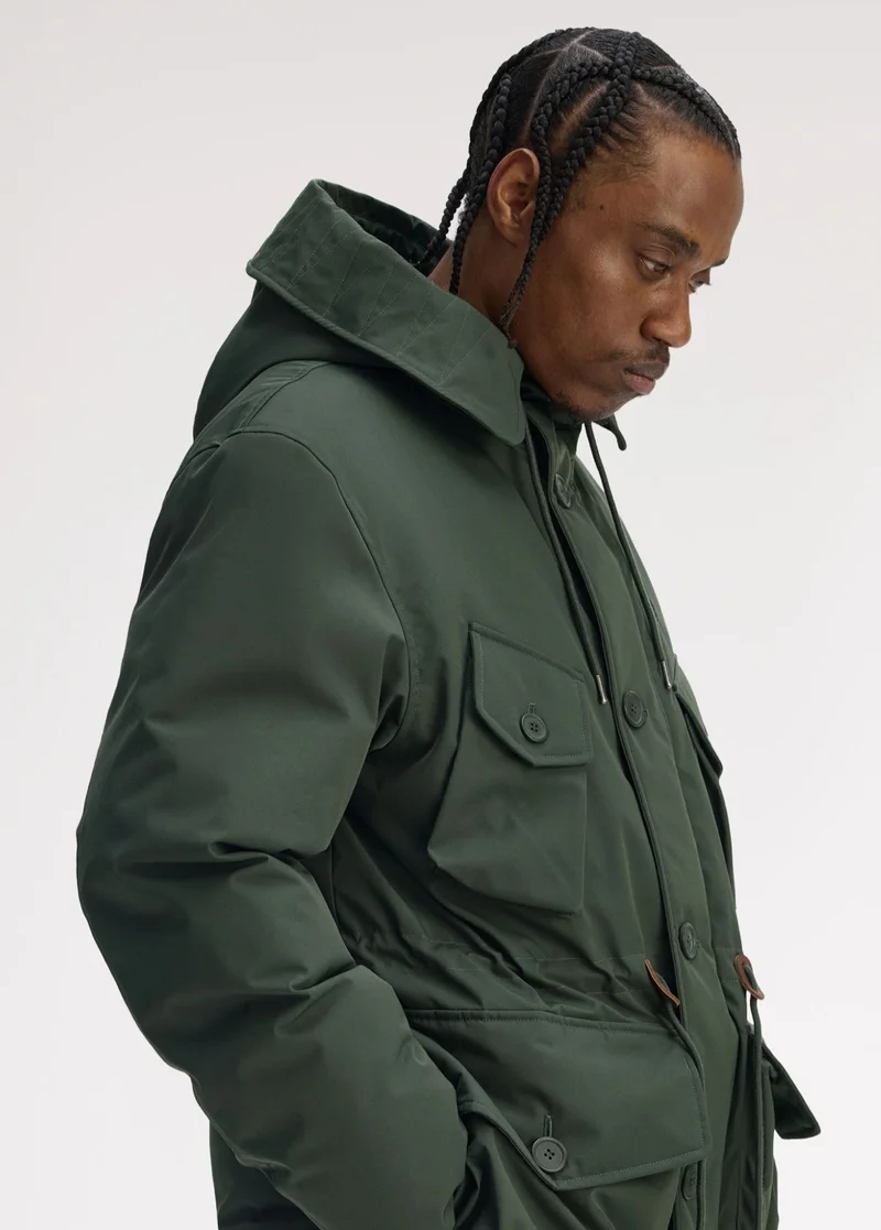 Black male model with braids stands to the side with his hands in the pockets of our heavyweight Patch Pocket Puffer Jacket in a natural dark green