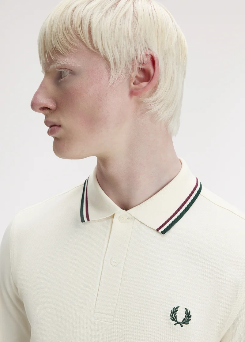White male model with with straight white hair and white eyebrows looks to his left and wears an Ecru Fred Perry Shirt with oxblood and dark green tipping