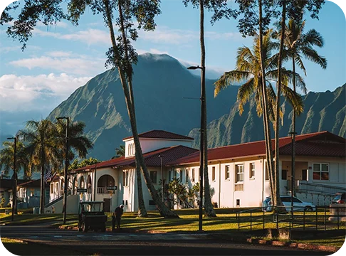 Image Description: A white building with a red tile roof surrounded by green mountains and palm trees.