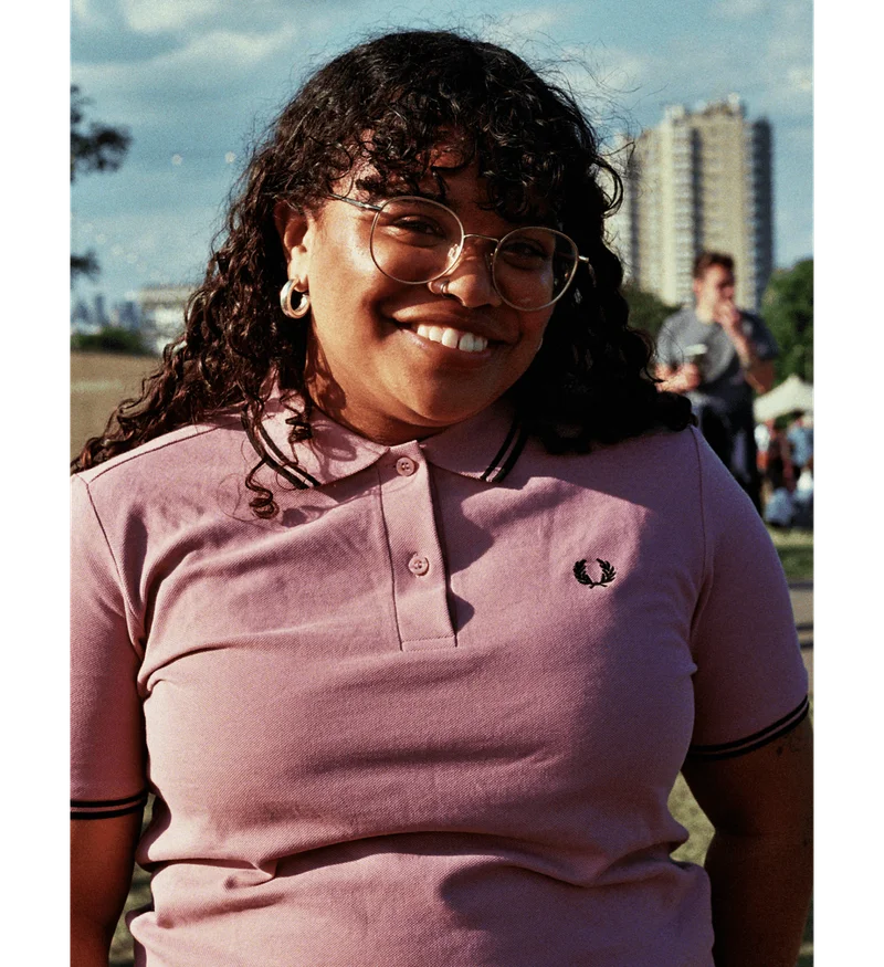 Woman smiling and sitting at a festival wearing a pink Fred Perry Shirt with black tipping