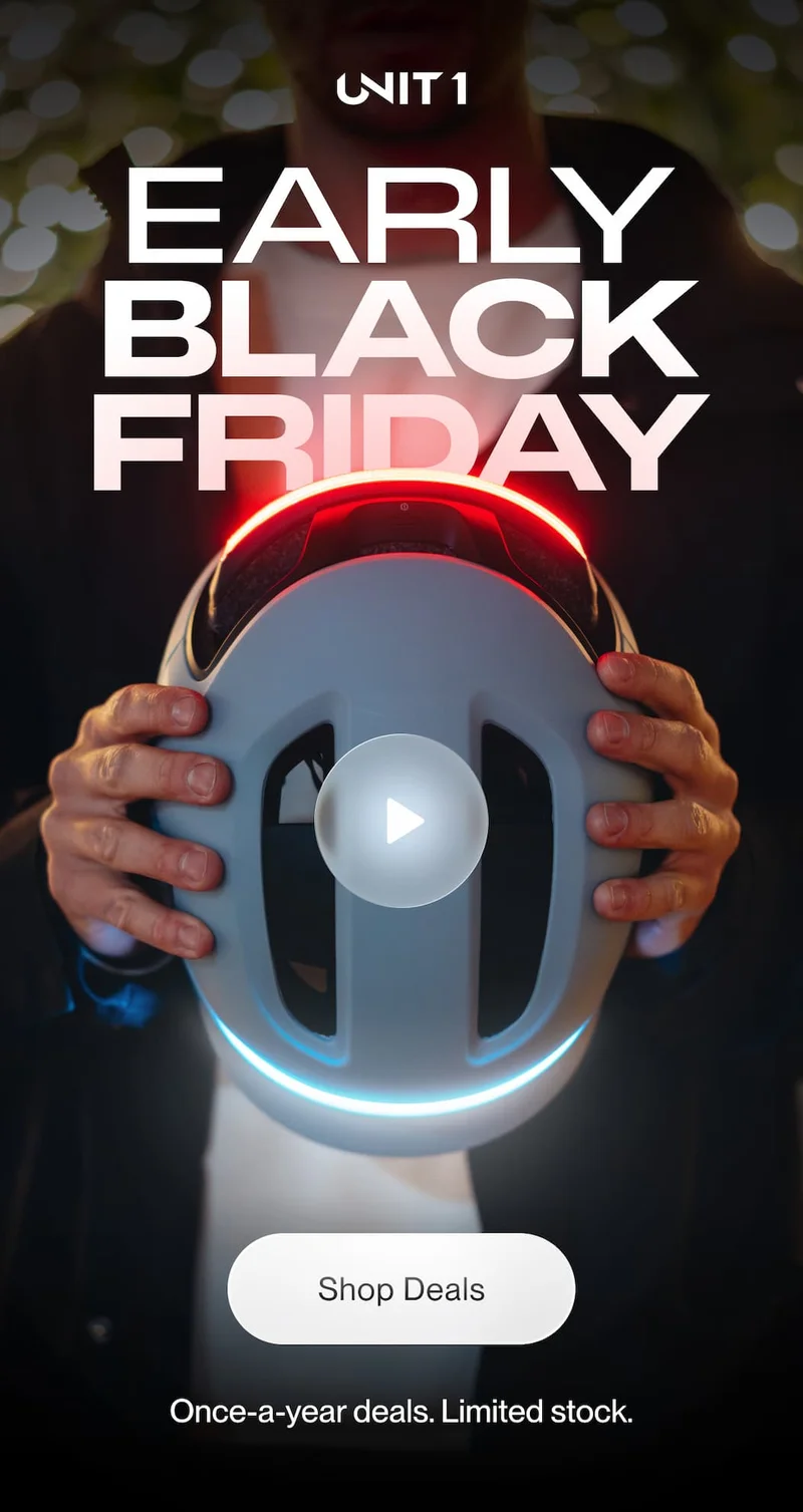 Person holding a futuristic smart bike helmet with glowing red and blue LED lights. Text above reads ‘UNIT 1 Early Black Friday’. Background softly blurred with warm bokeh lights.