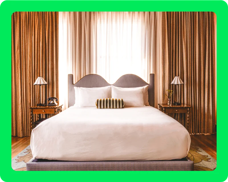 Image Description: Hotel room with white hotel bed in the center of the room. The bed has a headboard with a wavy shape and there are two simple wood nightstands next to either side of the bed. Behind the bed are heavy pale orange-colored curtains with light peeking through.