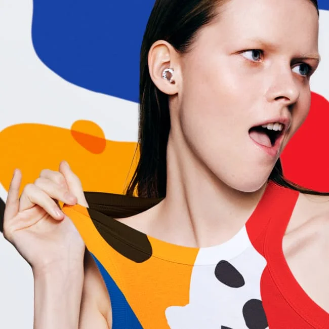 A woman wearing colorful Andrew Footit Experience earplugs tugs on a t-shirt that matches her earplugs.