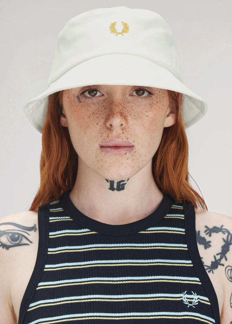 woman with ginger hair, freckles and tattoos stares into the camera wearing an ecru bucket hat and a black striped vest top