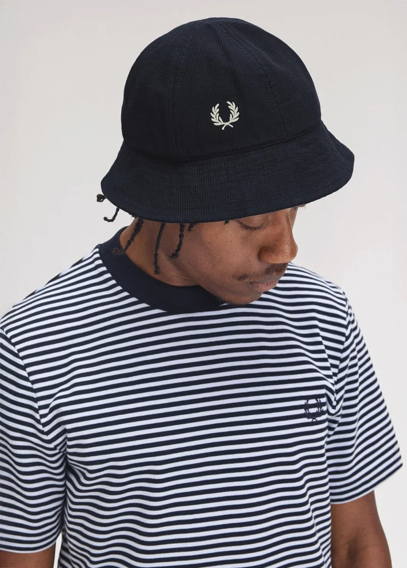 Black man looks towards the ground wearing a navy and white striped T-shirt and a navy seersucker bucket hat