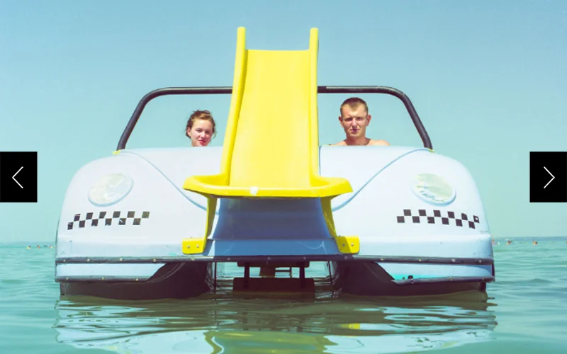 A man (right) and woman (left) are centered in a paddleboat amid shallow water and clear blue sky.