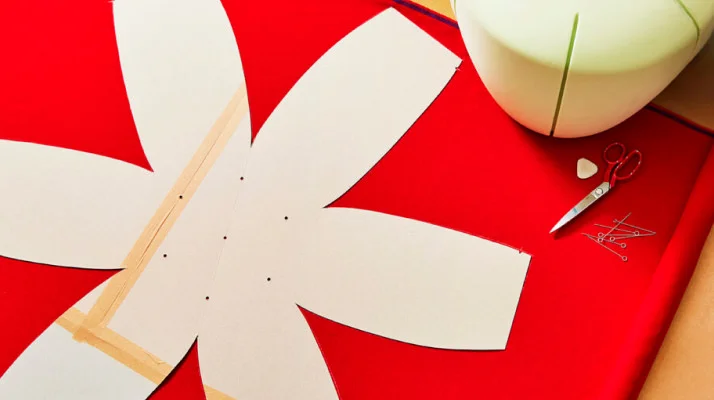 A flower shaped cutout template for the Truffle pouf's upholstery design sits over top of red fabric with scissors and the foam for the pouf in the top right corner.