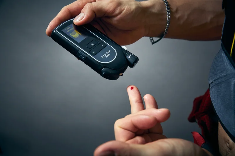 A close-up view of a finger with a drop of blood about to be applied to a glucose meter.