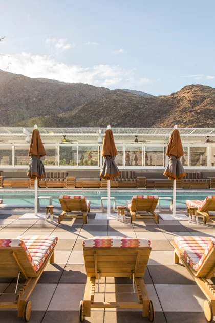 Cushioned wooden lounge chairs with umbrellas at a pool with lush mountains in view.