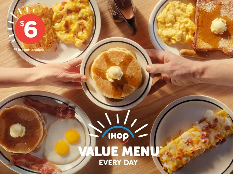 An assortment of IHOP Value Menu meals on a table, with two hands reaching across and holding a plate of pancakes topped with butter and syrup above it