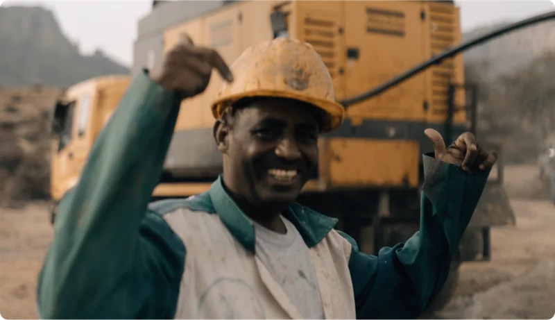 A man poses in front of a piece of construction equipment while pointing at the yellow hard hat he's wearing.