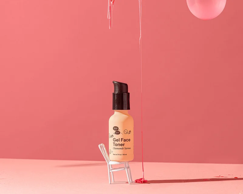 A light peach bottle sits on a miniature chair against a pink backdrop