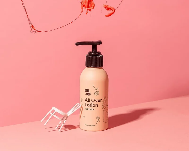 A light pink bottle with a toy chair tipped against it against a pink backdrop