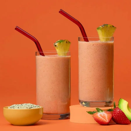 An  orange-themed setup featuring two fruit smoothies with a small bowl of seeds, fresh strawberries, and an avocado slice.