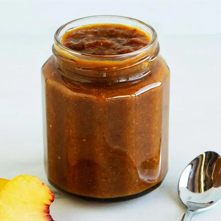 A hexagonal glass jar containing textured fruit jam, sitting on a white surface next to a metal spoon and a fresh peach slice.