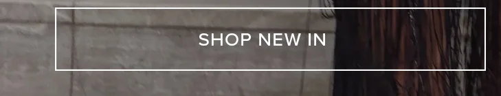 White "SHOP NEW IN" button on a blurred background featuring a grey textured wall and dark flowing hair. Discover the latest fashion arrivals.