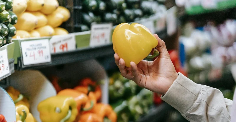 Hand holding a yellow bell pepper in a grocery store produce aisle with price signs visible