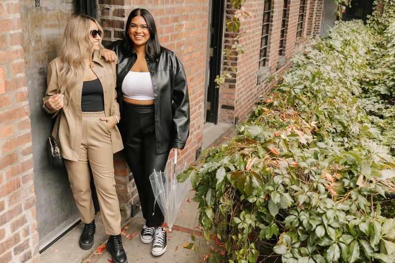 2 women wearing leather shirt and trousers stood in front of brick wall